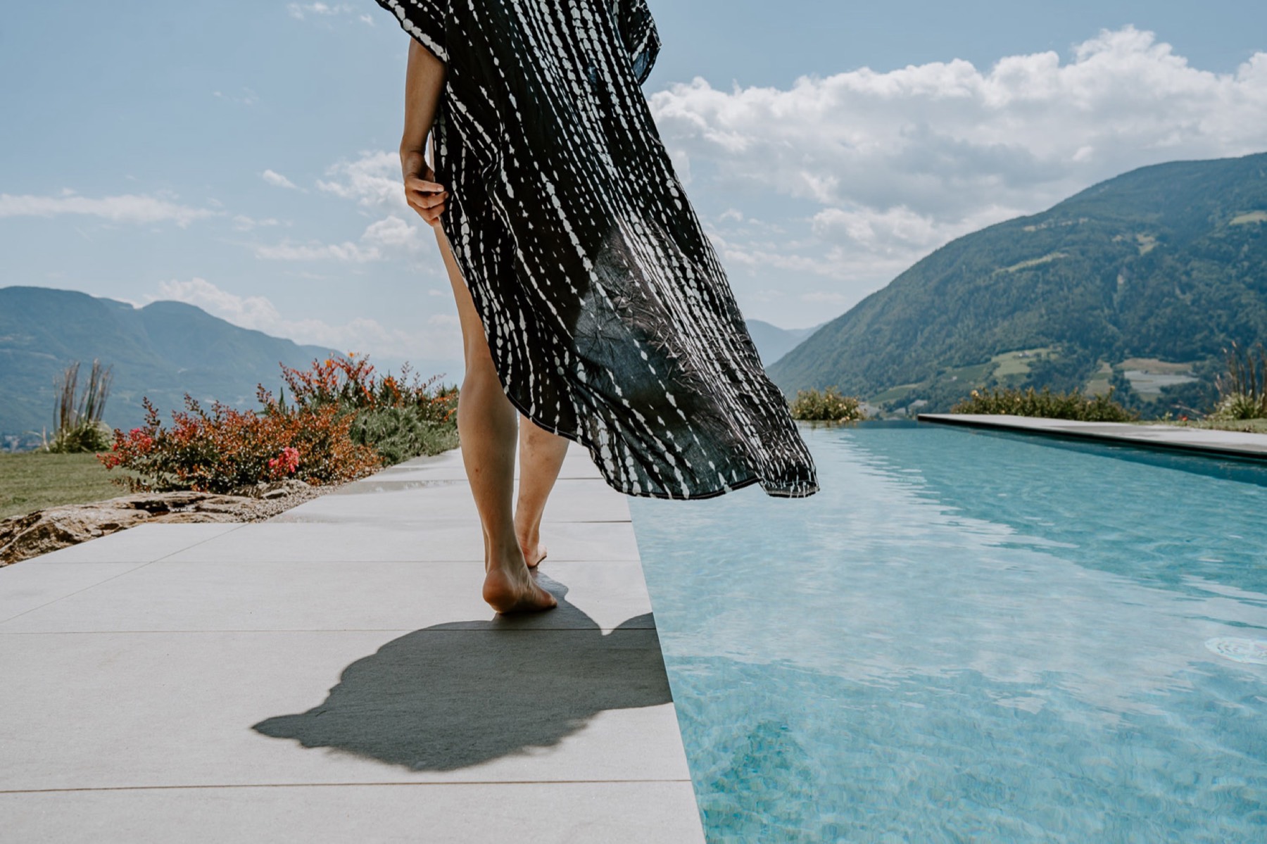 A guest in a flowing dress walking barefoot beside an infinity pool facing the mountains